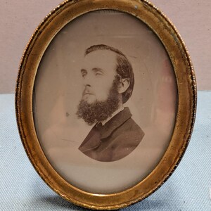 May include: An antique oval portrait of a man with a full beard, set within a gold-colored frame. The portrait is a sepia tone, showing the man in profile. The frame has a textured, ornate design.