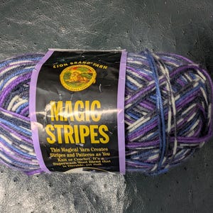May include: A skein of Lion Brand Magic Stripes yarn featuring a variegated color palette of purple, blue, white, and dark grey. The black paper label displays the brand logo, the text Magic Stripes, and descriptive product information.