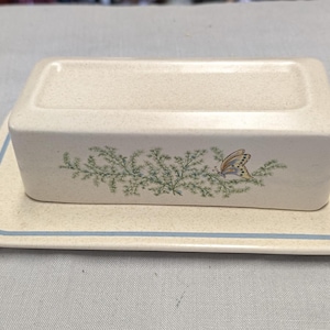 May include: A rectangular ceramic butter dish with a speckled off-white finish. The lid features a green botanical sprig design with a small butterfly. The base plate has a thin blue border line that frames the rectangular lid when placed on top.