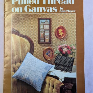 May include: A vintage book cover titled "Pulled Thread on Canvas" by Ann Meyer. The cover features a light blue embroidered pillow, framed needlework, a black patterned clutch, and a bouquet of flowers. The background is a patterned wallpaper.