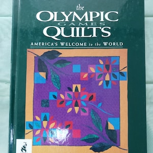 May include: A hardcover book titled "The Olympic Games Quilts" with the subtitle "America's Welcome to the World." The cover features a purple quilt design with floral and geometric patterns, framed in gold, and the Atlanta 1996 logo.