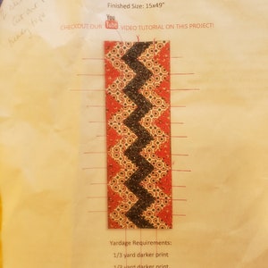 May include: A rectangular table runner pattern with a chevron design. The pattern features a black zigzag pattern on a red background with a floral design. The finished size is 15 x 49 inches.