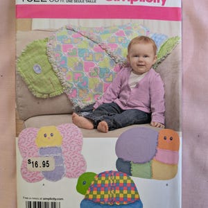 May include: Simplicity pattern 1822 featuring a baby sitting on a sofa with a colorful heart-patterned blanket. The pattern includes instructions for making a butterfly, snail, and turtle-shaped baby toys. The price is $16.95.