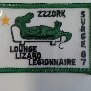 May include: Embroidered patch with a green border and a white background. The patch features a green alligator lounging on a chair, a gold star, and the text "ZZZORK LOUNGE LIZARD LEGIONNAIRE SURGE 87".