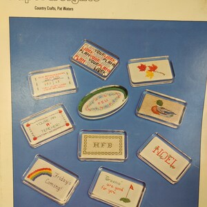 May include: A collection of paperweights with various cross-stitch designs. The paperweights feature phrases like "Plan Your Work" and "Friday's Coming!" along with images of a rainbow, a golf flag, and autumn leaves. The cover reads "Paperweights."