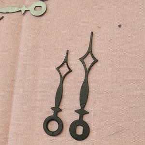 May include: Two black clock hands with diamond-shaped accents and circular bases. The hands are on a pink fabric surface.