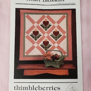 May include: A pattern for a wall and bed quilt titled "Heart Blossom" by thimbleberries. The quilt design features a diamond pattern with heart and floral motifs in shades of red, green, and cream. A wicker basket with dried flowers sits below the quilt.