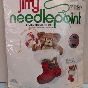 May include: A Jiffy Needlepoint kit titled "Jingles in a Stocking" featuring a Christmas ornament design. The kit includes a teddy bear holding a candy cane in a red stocking. The finished size is 5.5 inches high.