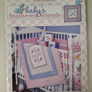 May include: A book cover titled "baby's feathered friends" featuring a crib with a pink and blue floral quilt and matching bedding. The cover has a pink and white checkered border and illustrations of birds.