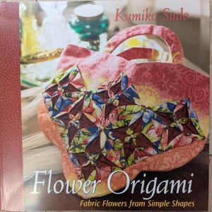 May include: A book titled Flower Origami by Kumiko Sudo featuring a fabric bag on the cover. The bag is decorated with folded fabric flower shapes in shades of red, blue, green, and yellow, accented with small white beads on a peach background.