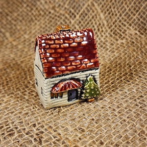 May include: A miniature ceramic house with a red-brown shingled roof, white walls, and a small green tree. The house has a small door and a decorative umbrella over the door. The house is a decorative item.