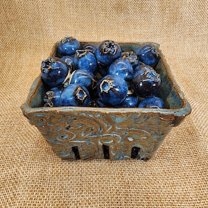 May include: A ceramic berry basket filled with glazed, dark blue ceramic blueberries. The basket is square with a brown and blue glaze and decorative floral patterns. The blueberries have a glossy finish and are arranged to fill the basket.