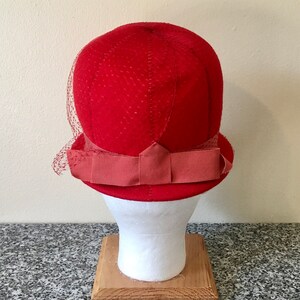 May include: A vintage red cloche hat with a small brim and a decorative bow. The hat features a fine mesh veil and is displayed on a white mannequin head. The hat is made of a textured fabric.