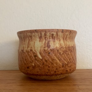 Pode incluir: Uma pequena xícara de cerâmica marrom e creme com um design texturizado ondulado. A xícara é feita à mão e tem um toque rústico e terroso.