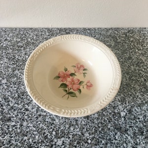 May include: Cream-coloured ceramic bowl with a floral design. The bowl has a decorative rim with a leaf pattern. The interior of the bowl displays a cluster of pink flowers with green leaves. A classic piece for serving.