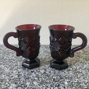 Könnte beinhalten: Zwei dunkelrote Glasbecher mit einem Fuß und einem dekorativen Design. Die Becher haben einen herzförmigen Griff.