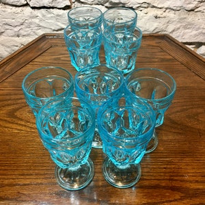 May include: Set of eight blue glass drinking glasses with a textured design. The glasses are arranged in a circle on a wooden table.