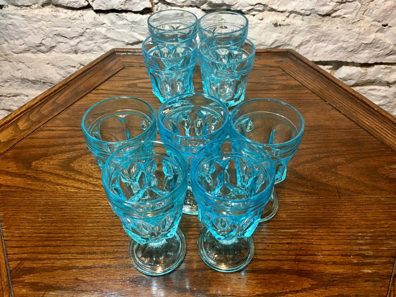 May include: Set of eight blue glass drinking glasses with a textured design. The glasses are arranged in a circle on a wooden table.