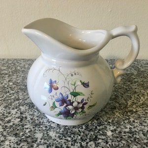 Puede incluir: Jarra de cerámica blanca con cuerpo redondeado y asa curva. La jarra está decorada con un diseño floral con flores moradas y azules y hojas verdes. La jarra tiene un pico para verter fácilmente.