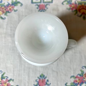 May include: White ceramic cup with a rounded bottom and a handle. The cup is empty and appears to be vintage.
