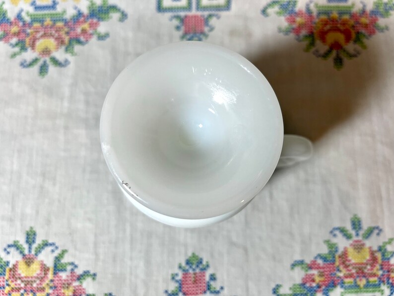 May include: White ceramic cup with a rounded bottom and a handle. The cup is empty and appears to be vintage.