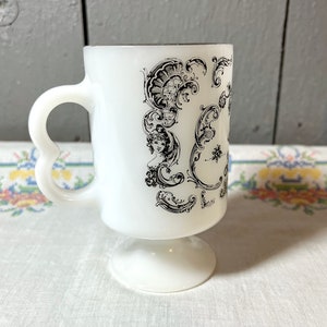 May include: White ceramic mug with a black and white floral design. The mug has a heart-shaped handle and a pedestal base.