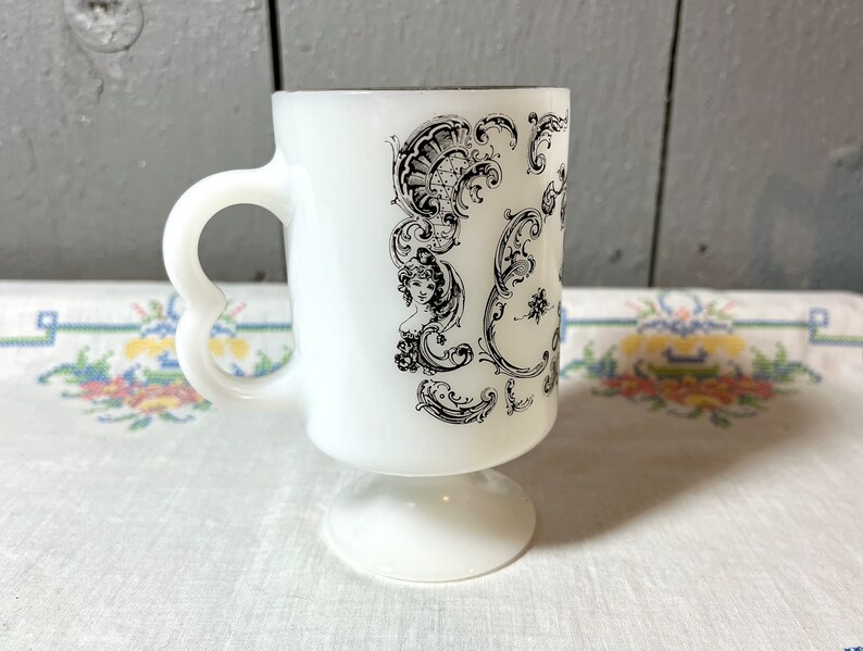 May include: White ceramic mug with a black and white floral design. The mug has a heart-shaped handle and a pedestal base.
