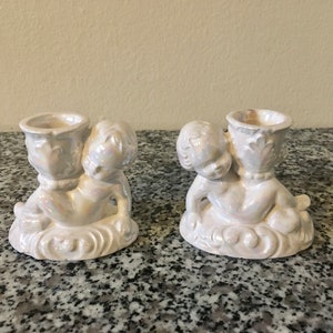 Pode incluir: Dois castiçais de querubim brancos perolados. Cada querubim está ajoelhado e segurando uma taça de vela decorativa. Os castiçais estão sobre uma base branca em forma de nuvem. Os castiçais são feitos de cerâmica.