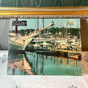 May include: A 1000 piece jigsaw puzzle box featuring a colourful image of sailboats docked in a harbour. The box is brown with white text that reads "1000 piece jigsaw puzzle".