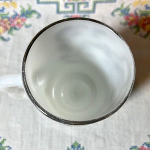 May include: A white milk glass cup with a silver rim. The cup is empty and has a slight swirl pattern on the inside.