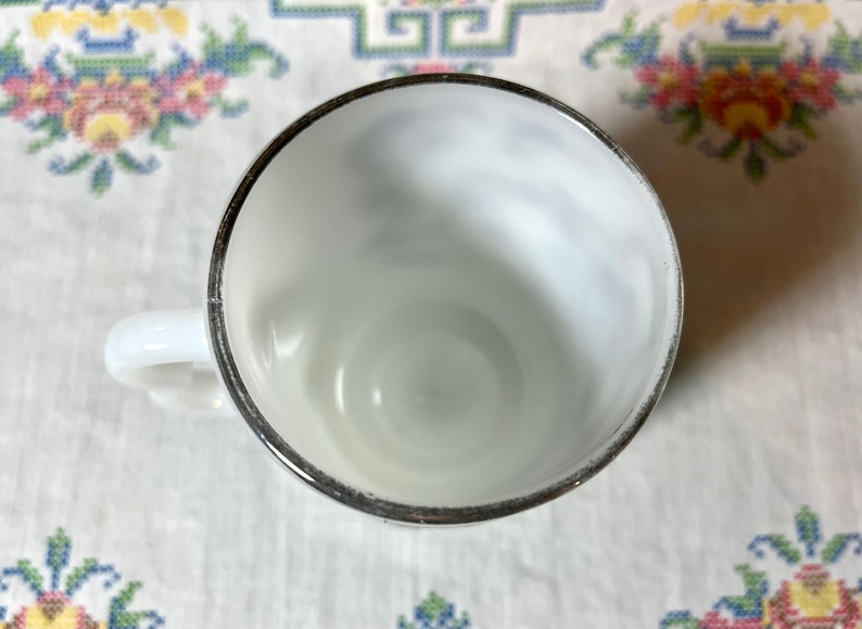 May include: A white milk glass cup with a silver rim. The cup is empty and has a slight swirl pattern on the inside.