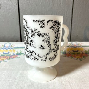 May include: White ceramic mug with a black and white design of cherubs and floral patterns. The mug has a handle and a pedestal base. The text "Cup" is visible on the mug.