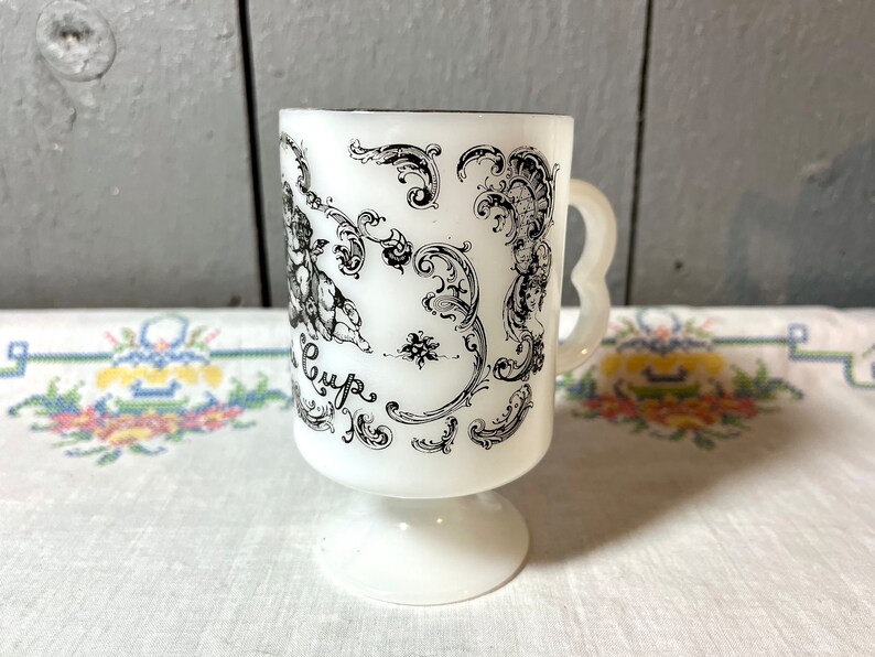 May include: White ceramic mug with a black and white design of cherubs and floral patterns. The mug has a handle and a pedestal base. The text "Cup" is visible on the mug.