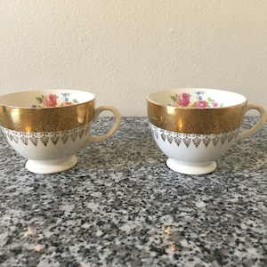 Op de afbeelding: Twee witte theekopjes met gouden rand en bloemenpatronen. De kopjes hebben een gouden band rond de bovenkant en een gouden bloemenontwerp onder de band.