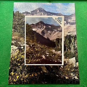 May include: A 500 piece jigsaw puzzle depicting a mountain scene with a blue sky and white clouds. The puzzle is titled "Rocky Mountains" and is made by "Interlocking" with the number "500" printed on the box.
