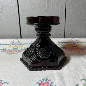 Puede incluir: Un portavelas de vidrio negro con un diseño decorativo. El portavelas tiene una parte superior redonda y una base cuadrada.