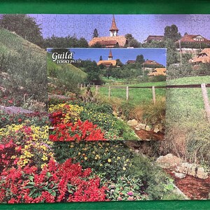 May include: A colourful jigsaw puzzle depicting a small village with a church in the background. The puzzle is 1000 pieces and is made by Guild.