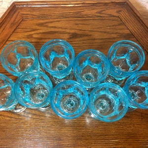 May include: A set of nine blue glass drinking glasses with a textured design. The glasses are arranged in a circle on a wooden surface.