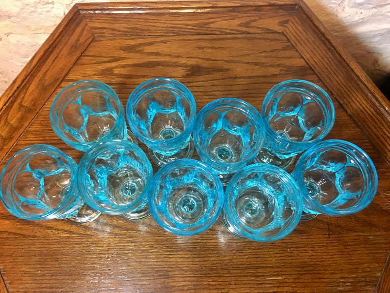 May include: A set of nine blue glass drinking glasses with a textured design. The glasses are arranged in a circle on a wooden surface.
