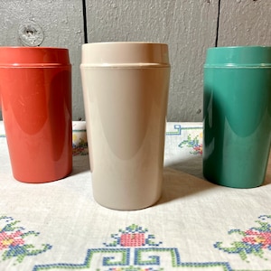 Könnte beinhalten: Drei Plastikbecher in verschiedenen Farben: Koralle, Beige und Türkis. Die Becher stehen auf einer weißen Tischdecke mit Blumenmuster.