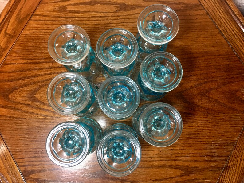 May include: A set of ten blue glass cups with a textured design. The cups are arranged in a circle on a wooden surface.