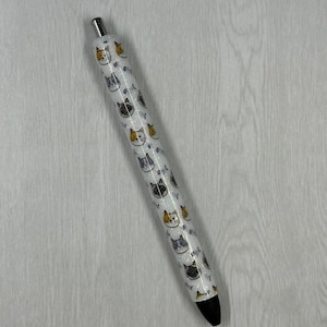 May include: A white ballpoint pen with a repeating pattern of cat faces in orange, gray, and black. The pen has a silver tip and a black ink cartridge. The pen is approximately 5.5 inches long.