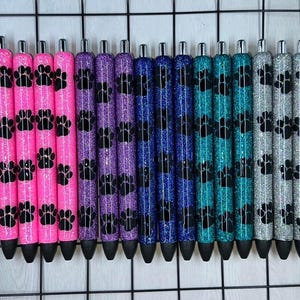 May include: A set of 15 glitter pens with black paw print designs. The pens are in various colors, including pink, purple, blue, teal, and silver.