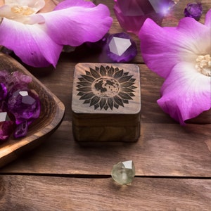 Peut inclure: Une boîte en bois avec un motif de tournesol gravé sur le couvercle. Le tournesol a une lune au centre. La boîte est entourée de fleurs violettes et de cristaux.