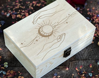 May include: A wooden box with a light wood finish and a gold engraved design of two hands holding a sun and moon. The box has a brass latch.
