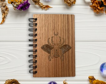 May include: A wooden spiral bound journal with a laser engraved design of a beetle with wings and a crescent moon. The journal is on a white wooden surface with dried flowers and blue glass beads.