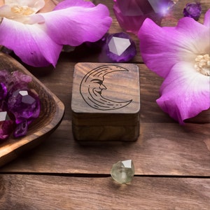 Peut inclure: Une boîte en bois avec un motif de croissant de lune gravé sur le couvercle. La boîte est entourée de cristaux violets et verts et de fleurs roses.