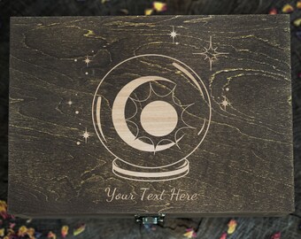 May include: A dark brown wooden box with a hinged lid. The box has a design of a crystal ball with a crescent moon and stars engraved on the front. The text "Your Text Here" is engraved below the design.