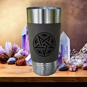 Op de afbeelding: Een roestvrijstalen tumbler met een grijze leren omhulsel. De omhulsel heeft een zwart pentagram met schedels op elk punt.