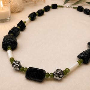 May include: A necklace featuring irregular black stone beads, smooth white cylindrical spacers, small round olive green beads, and two metallic silver skull-shaped beads arranged in a repeating pattern along the length of the jewellery piece.